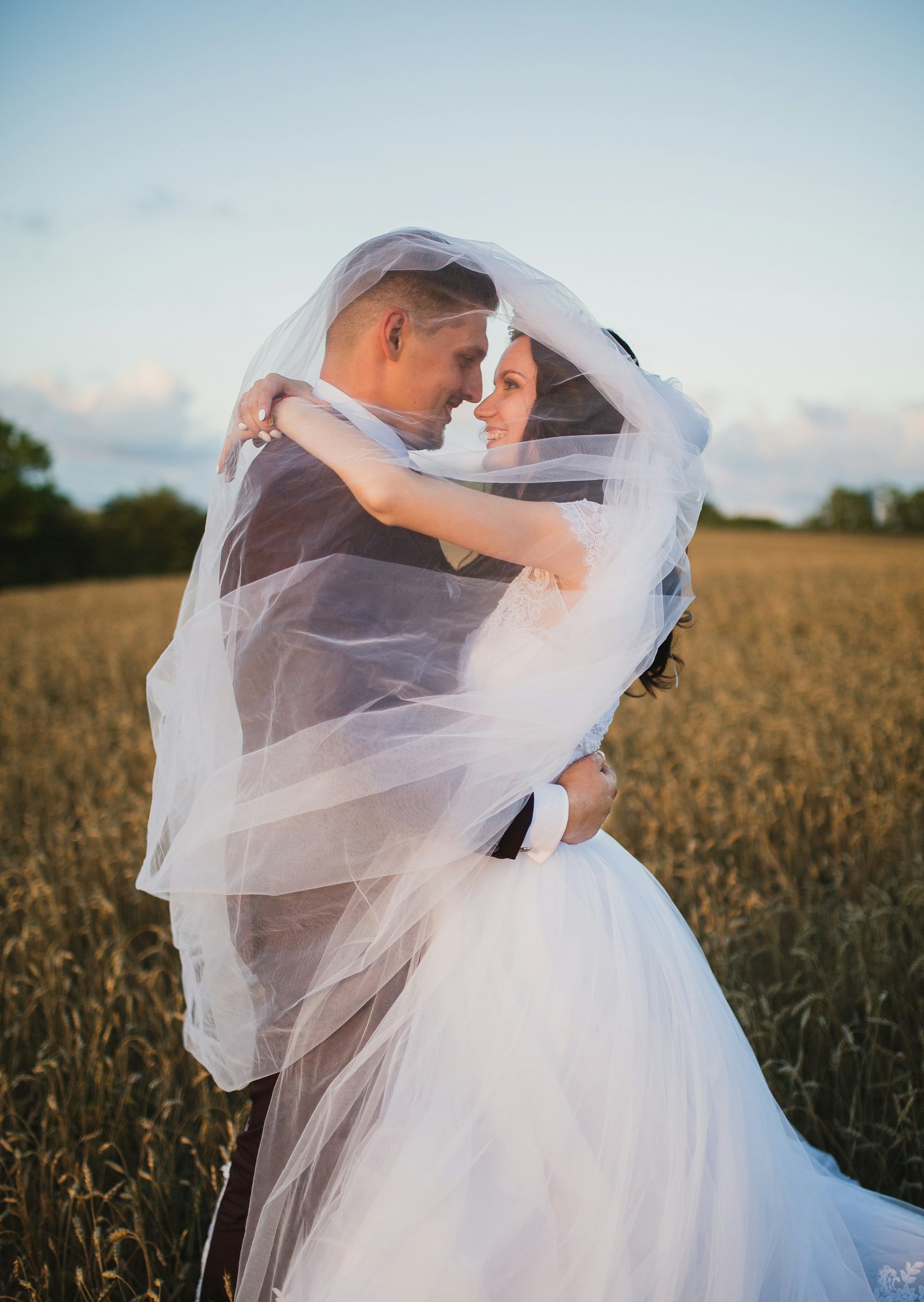 Bride and groom embracing under a veil in a golden wheat field at sunset